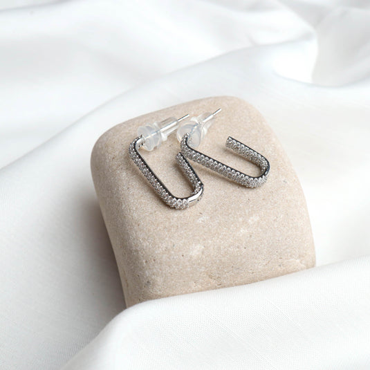 QuinShine sparkling silver elongated hoop earrings for women on a beige stone with white fabric background