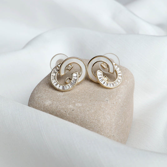 QuinShine Lunar Link gold earrings with clear crystals on a soft beige stone and white fabric background
