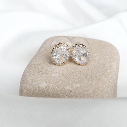 QuinShine aura oval stud earrings with clear gemstones and gold settings displayed on a beige stone