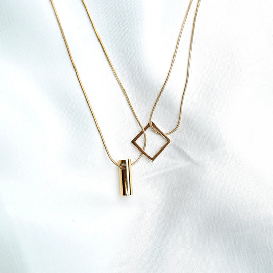 Gold cylindrical bar pendant necklace with a thin chain displayed on white fabric background
