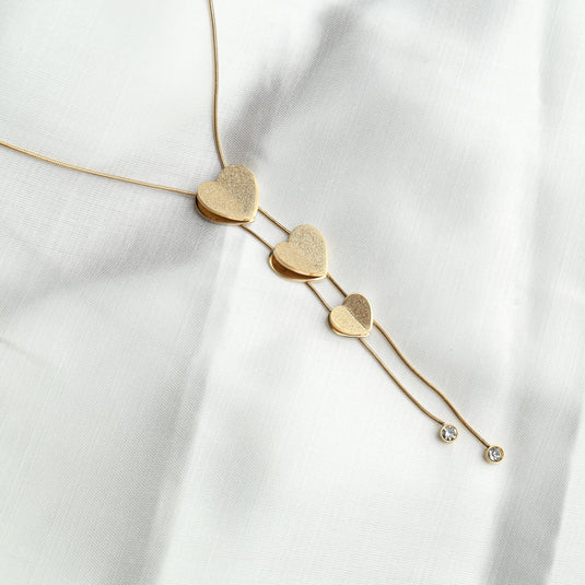 Elegant gold heart lariat necklace with three textured heart pendants and crystal accents on a white fabric
