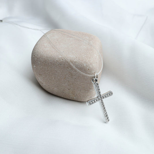 QuinShine silver cross necklace with sparkling stones displayed on a beige stone and white fabric background