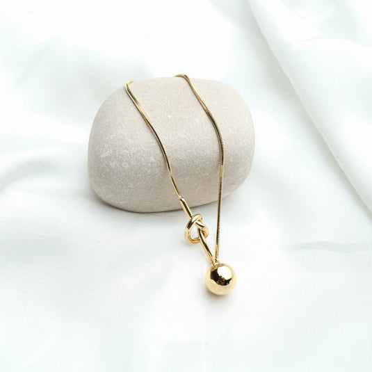 QuinShine gold knot lariat necklace with polished ball pendant displayed on a smooth stone